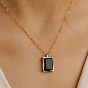 May include: Gold-toned necklace with a rectangular black pendant. The pendant is set in a gold frame and hangs from a delicate gold chain. The necklace is displayed on a person's neck, showcasing its elegant design.