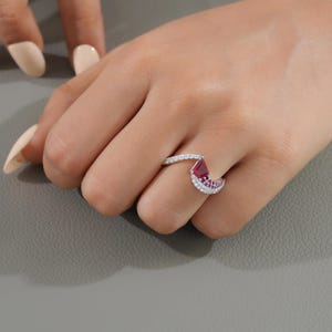 May include: A silver ring with a pear-shaped, deep red gemstone and a curved band. The band is encrusted with small, clear gemstones. The ring is on a hand with manicured nails. The logo "MANSI JEWELS" is at the bottom.