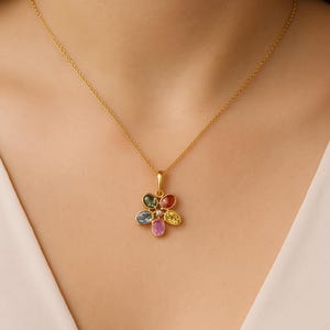 Op de afbeelding: Een goudkleurige ketting met een bloemhanger met kleurrijke edelstenen. De bloem heeft zes bloemblaadjes, elk in een andere kleur: blauw, groen, rood, geel en roze. In het midden zit een kleine diamant. De ketting wordt gedragen.