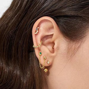 Może przedstawiać: Zbliżenie ucha ozdobionego wieloma złotymi kolczykami. Ucho posiada kolczyk helix, kolczyk łańcuszkowy z zielonymi i złotymi akcentami oraz kolczyk sztyft z małym diamentem.