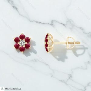 Puede incluir: Un par de pendientes de oro con cierre de tuerca. Cada pendiente presenta un diseño floral con seis gemas rojas que rodean una gema blanca central. Los pendientes se exhiben sobre una superficie de mármol blanco.