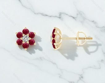 May include: A pair of gold stud earrings. Each earring features a flower design with six red gemstones surrounding a central white gemstone. The earrings are displayed on a white marble surface.