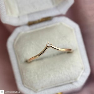 Pode incluir: Um delicado anel dourado com um design em forma de V, apresentando um pequeno diamante brilhante no ápice. O anel é apresentado numa caixa de joias branca com um interior macio e aveludado. É uma peça de joalharia fina.