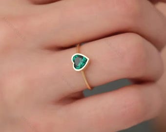 May include: A gold ring featuring a heart-shaped, emerald-green gemstone. The gemstone is set in a white bezel, and the ring band is a thin, polished gold. The ring is shown on a person's finger.