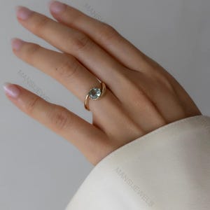 May include: A gold ring featuring a light blue gemstone. The ring's design is a distinctive swirl that holds the oval-shaped stone. The ring is displayed on a hand, with a white sleeve visible in the background.