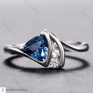 Puede incluir: Un anillo de plata con una gran piedra preciosa triangular de color azul oscuro. El anillo tiene un diseño moderno con una banda curva y pequeñas piedras de acento redondas y claras. El anillo está sobre un fondo gris.