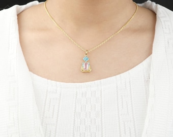 May include: A gold-colored necklace with a pendant featuring a light blue and pink gemstone design. The pendant is rectangular with a smaller, rounder stone above it. The necklace is worn against a white background.