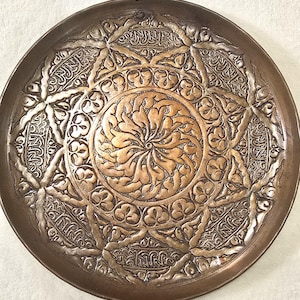 May include: A round, antique copper-colored metal tray with intricate embossed floral and geometric patterns. The center features a stylized floral design, surrounded by detailed arabesque motifs and script. The tray's rim has a darker, aged patina.
