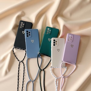 May include: A collection of colorful phone cases in black, blue, green, white, and lavender. Each case features a heart design and a matching lanyard. The cases are displayed on a beige fabric background.