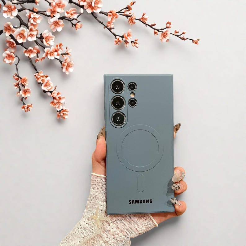 Peut inclure: Coque de t&eacute;l&eacute;phone bleu ardoise avec un design circulaire et le logo Samsung. L'&eacute;tui poss&egrave;de un module cam&eacute;ra avec plusieurs objectifs. L'&eacute;tui est tenu dans une main avec une manche en dentelle, avec un fond de fleurs roses.