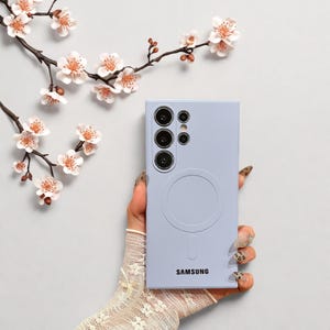 Peut inclure: Une coque de t&eacute;l&eacute;phone Samsung violet clair avec un design circulaire et le nom de la marque imprim&eacute; en bas. L'&eacute;tui comporte plusieurs d&eacute;coupes pour l'appareil photo. Le fond comprend un arrangement floral avec des fleurs blanches et roses.