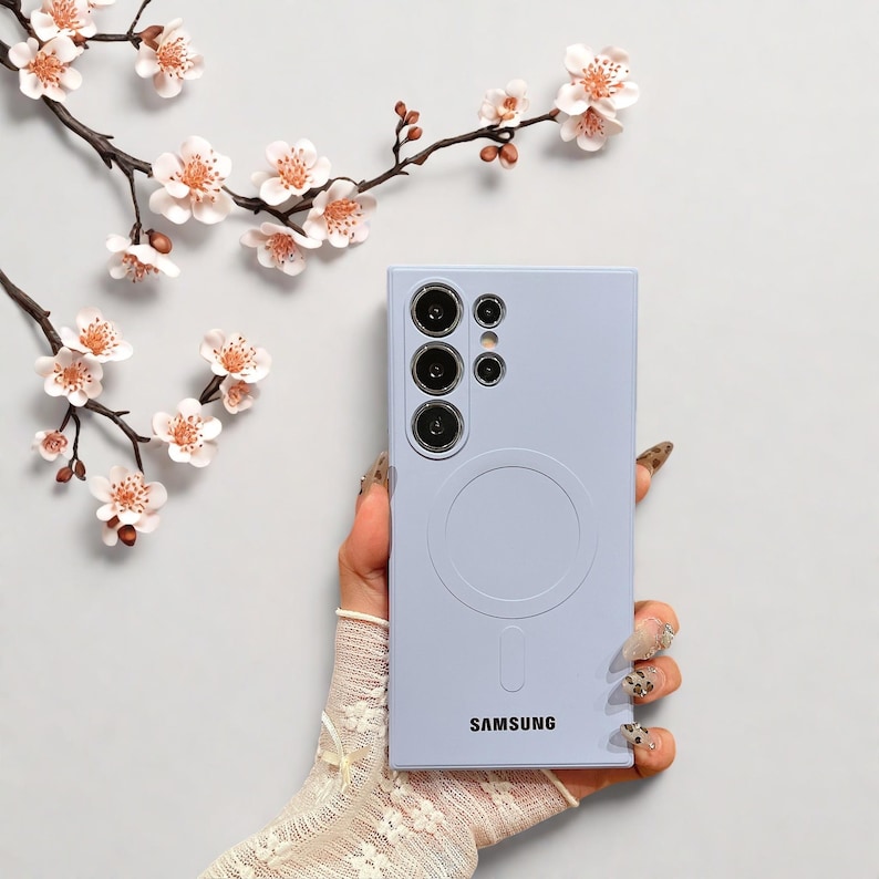 Peut inclure: Une coque de t&eacute;l&eacute;phone Samsung violet clair avec un design circulaire et le nom de la marque imprim&eacute; en bas. L'&eacute;tui comporte plusieurs d&eacute;coupes pour l'appareil photo. Le fond comprend un arrangement floral avec des fleurs blanches et roses.