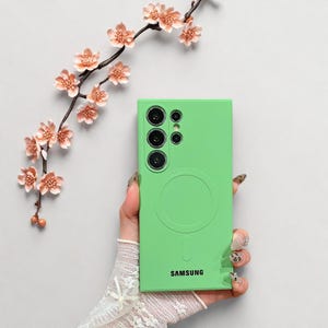 Peut inclure: Coque de t&eacute;l&eacute;phone vert vif avec un design circulaire et le mot "SAMSUNG" en bas. L'&eacute;tui pr&eacute;sente une d&eacute;coupe pour l'appareil photo avec plusieurs objectifs. Une branche florale d&eacute;corative avec des fleurs roses en arri&egrave;re-plan.