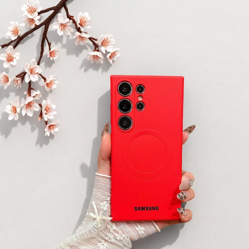 Peut inclure: Une coque de t&eacute;l&eacute;phone rouge vif avec un design circulaire et le mot "SAMSUNG" imprim&eacute; en bas. L'&eacute;tui comporte plusieurs d&eacute;coupes pour l'appareil photo. En arri&egrave;re-plan, une branche avec des fleurs blanches.