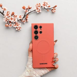 Peut inclure: Une coque de t&eacute;l&eacute;phone corail avec un design circulaire et le logo Samsung. L'&eacute;tui comporte plusieurs d&eacute;coupes pour l'appareil photo et un cercle de chargement magn&eacute;tique. Des fleurs d&eacute;coratives sont en arri&egrave;re-plan.