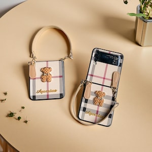 May include: Two beige and black plaid phone cases with a teddy bear design and the word "Aquarius". One case has a handle, the other has a strap. The cases are on a light brown surface.