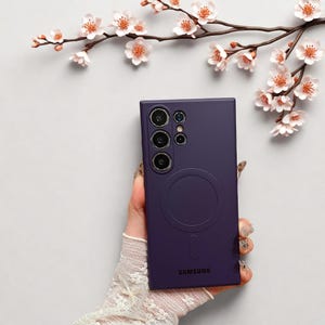 Peut inclure: Une coque de t&eacute;l&eacute;phone violet fonc&eacute; avec un design circulaire et le logo Samsung. L'&eacute;tui a une forme rectangulaire avec plusieurs d&eacute;coupes pour l'appareil photo. Le fond pr&eacute;sente une surface blanche et une branche avec des fleurs blanches et roses.