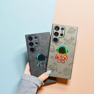 May include: Two phone cases with space-themed designs. One case is dark gray, the other is a lighter gray. Each case features an astronaut with a rainbow-colored visor and space-related illustrations. The cases are held against a blue and peach background.