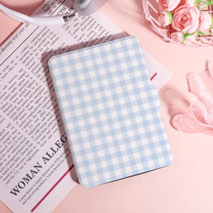 May include: A notebook or tablet cover with a light blue and white gingham pattern. The cover is rectangular and has a soft, pastel colour scheme. The background is a light pink surface with a newspaper and flowers.