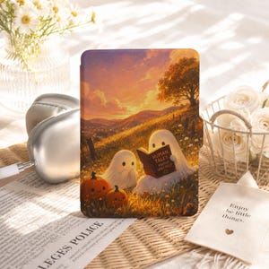 May include: A rectangular e-reader case featuring a printed illustration of two white ghosts sitting in a grassy field at sunset. One ghost holds an open book titled Human Tales Mostly Very Silly. Two small orange pumpkins with faces sit nearby.