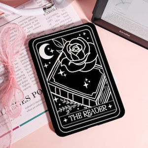 May include: Black rectangular phone case with a white illustration of a rose, moon, stars, and a book. The text "THE READER" is at the bottom. The case is on a pink surface.