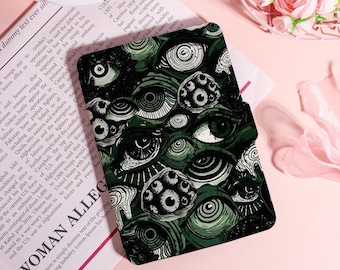 May include: Dark green case with a repeating pattern of stylized eyes in various sizes and styles. The case is on a pink surface with a newspaper and a pink ribbon in the background. The text "WOMAN ALLEG" is visible.