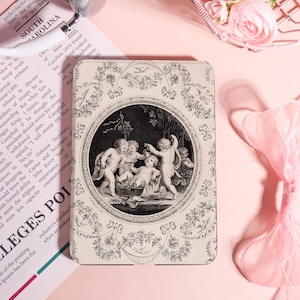 Könnte beinhalten: Dekorative, rechteckige Hülle mit einer Schwarz-Weiß-Illustration von Engeln in einem kreisförmigen Rahmen. Das Gehäuse hat ein florales Bordürendesign auf rosa Hintergrund.