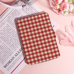 May include: A red and white checked phone case. The case has a classic gingham design. The image also shows a newspaper and text that reads "WOMAN ALLEG".