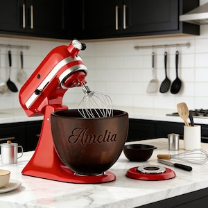 Puede incluir: Una batidora de pie roja con un batidor de acero inoxidable y un cuenco de madera. El cuenco está grabado con el nombre "Amelia". Hay utensilios de cocina adicionales en la encimera, como un cuenco pequeño, una base roja y un batidor.