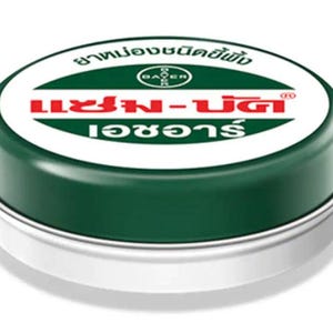 Könnte beinhalten: Eine runde, grüne Dose mit einem weißen Etikett. Das Etikett trägt den Text "Bayer" in einem roten Kreis und den Text "Mentholatum" in roten Buchstaben. Der Text "อย่าทิ้งขยะลงแม่น้ำ" ist in schwarzen Buchstaben.