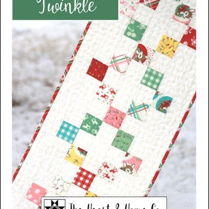 Puede incluir: Un camino de mesa de tela blanca con el título "Twinkle" y un patrón de cuadrados rojos, verdes y amarillos con temática navideña. La tela está acolchada y tiene el logotipo "The Heart & Home Co." en la parte inferior.