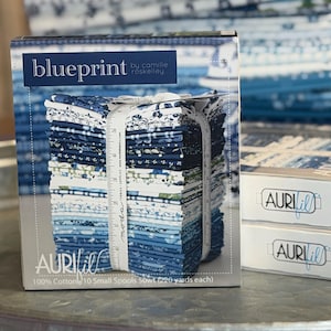 May include: A rectangular box labeled Blueprint by Camille Roskelley containing a stack of folded fabric squares in shades of blue and white. A white measuring tape is wrapped around the fabric stack, showing measurements up to 6 inches.
