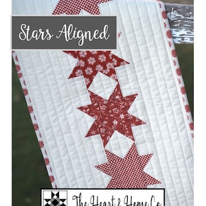 May include: A white table runner with a red and white star pattern. The runner has a red and white gingham border. The text "Stars Aligned" is printed on the runner. The text "The Heart & Home Co." is printed on the runner.