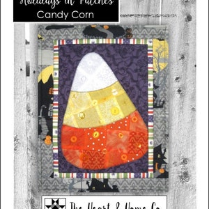 Może przedstawiać: Blok patchworkowej kołdry przedstawiający projekt w kształcie cukrowego kukurydzy w kolorystyce białej, żółtej i pomarańczowej. Blok jest otoczony purpurową tkaniną z kwiatowym wzorem i czarno-biało-czerwoną paskowaną obwódką. Blok kołdry nosi tytuł "Holidays in Patches Candy Corn" i jest oznaczony jako "HNH16". Blok kołdry został wykonany przez The Heart & Home Co.