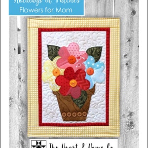Puede incluir: Un tapiz de pared acolchado con un diseño floral. Las flores están hechas de tela y tienen botones para centros. Las flores están en una maceta marrón con un fondo a cuadros amarillo y blanco. El texto "Holidays in Patches Flowers for Mom" está en la parte superior de la imagen. El texto "The Heart & Home Co." está en la parte inferior de la imagen.