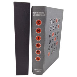 May include: A grey hardback book, "Enigma" by Hugh Sebago-Montefiore, with a black slipcase. The book's spine displays the title's letters in red circles. The Folio Society logo is at the bottom. The book is in the English language.