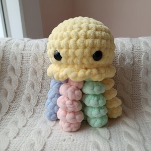 May include: A handmade crochet octopus plush toy. The octopus has a yellow head with black eyes and a ruffled edge. Its eight tentacles are pastel colors: blue, pink, green, and yellow. The plush is soft and sits on a white cable-knit blanket.