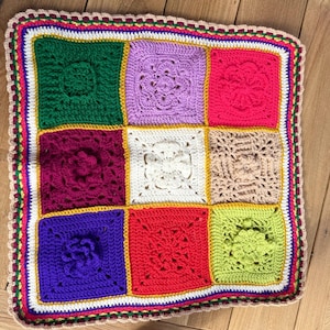 May include: A colourful crocheted square blanket with a variety of patterns and colours, including green, purple, red, white, and brown. The blanket is bordered with a multi-coloured trim.