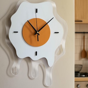 以下が含まれることがあります： 目玉焼きの形をした、気まぐれな壁掛け時計。時計は、透明なアクリルボーダー、オレンジ色の黄身、黒い針が付いた白い卵白のデザインが特徴です。明るいベージュの壁に取り付けられています。