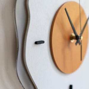 May include: A contemporary wall clock featuring a white, undulating frame and a light brown circular centre. The clock hands are black, and the hour markers are small, black rectangles. The clock is mounted on a white wall.
