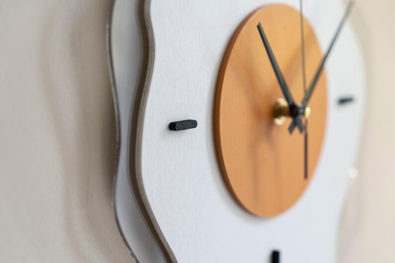 May include: A contemporary wall clock featuring a white, undulating frame and a light brown circular centre. The clock hands are black, and the hour markers are small, black rectangles. The clock is mounted on a white wall.