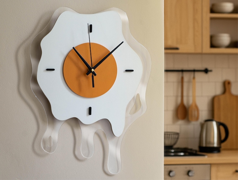 May include: A quirky wall clock in the shape of a fried egg. The clock has a white egg white design with a clear acrylic border, an orange yolk, and black hands. It is mounted on a light beige wall.