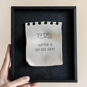 May include: A black framed ceramic notepad with the words "TO DO: -write a to do list!" written on it. The notepad is white with a torn edge and a series of holes at the top. The artwork is held by a person.