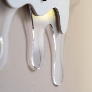 May include: A contemporary wall clock with a distinctive design. The clock has a white wooden base with clear, acrylic, drip-shaped accents. The clock's design is minimalist and artistic, adding a modern touch to any room.