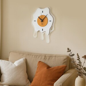 May include: A quirky wall clock in the shape of a fried egg. The clock has a white 'egg white' design with a dripping effect, an orange 'yolk', and black clock hands. It's mounted on a beige wall above a beige sofa.
