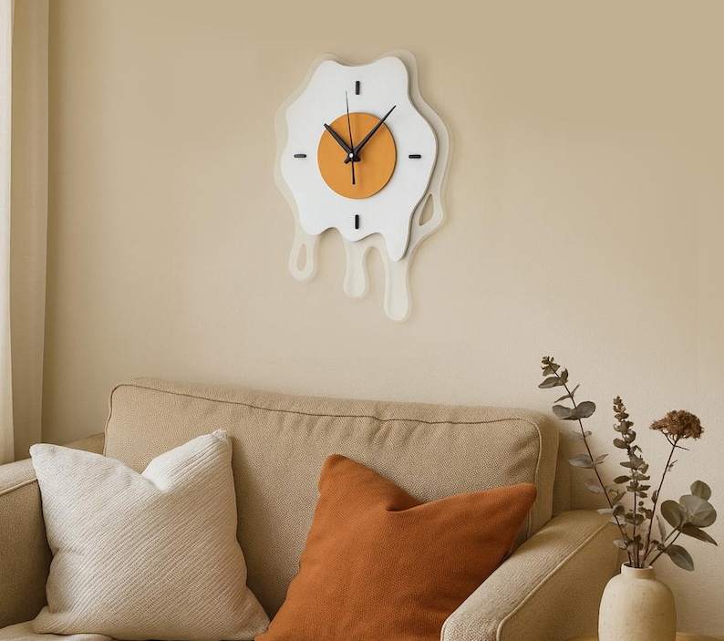 May include: A quirky wall clock in the shape of a fried egg. The clock has a white 'egg white' design with a dripping effect, an orange 'yolk', and black clock hands. It's mounted on a beige wall above a beige sofa.
