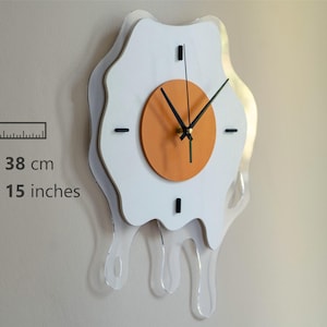 May include: A quirky wall clock in the shape of a fried egg. The clock has a white egg outline, an orange clock face, and black hands. It measures 28 x 38 cm and features a clear, dripping acrylic design.