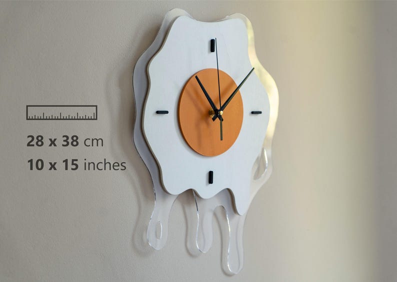 May include: A quirky wall clock in the shape of a fried egg. The clock has a white egg outline, an orange clock face, and black hands. It measures 28 x 38 cm and features a clear, dripping acrylic design.