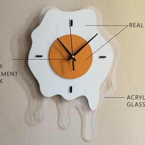 May include: A quirky wall clock in the shape of a fried egg, with a white egg white and an orange yolk. It has black hands and markers, made from real wood and acrylic glass. The text "QUIET SWEEP MOVEMENT CLOCK" is displayed on the left.