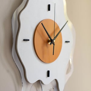 May include: A quirky wall clock shaped like a fried egg. It has a white, irregular border, a light brown central circle, and black clock hands. The clock's dimensions are not visible in the image.